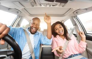 We Are Going On Vacation. Frontal view portrait of cheerful positive black guy and lady driving their automobile, dancing to music and singing favorite song, two people looking at camera photo