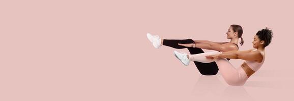 Two women engage in synchronized core exercises on a pink backdrop, demonstrating flexibility and strength. Both are focused and determined while showcasing proper form. photo