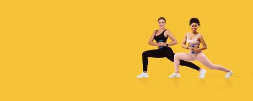 Two women engage in a lunge exercise while holding dumbbells. They are dressed in athletic wear and display focused expressions, set against a vibrant yellow background. photo