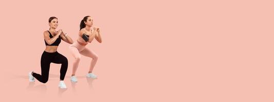 Two women are engaged in a squat exercise, showcasing different workout outfits. They are focused and demonstrating proper form on a solid pink backdrop during a fitness session. photo