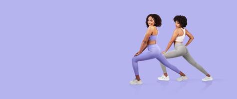 Two women are performing lunges in stylish activewear against a soft purple backdrop. Their focused expressions highlight dedication to fitness and healthy living. photo