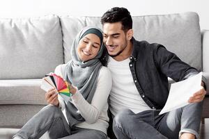 Happy middle-eastern family working on interior for their new house. Cheerful arabic man and woman in hijab sitting on floor by paper boxes with color samples, choosing colors and textures, copy space photo
