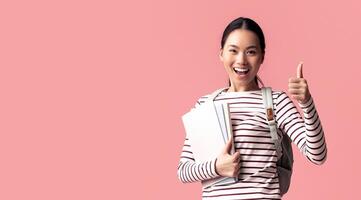 Cheerful Asian Female Student Jumping In Air With Workbooks And Showing Thumb Up, Happy Excited Korean Female Wearing Backpack And Smiling At Camera While Posing Over Pink Background, Copy Space photo