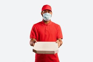 Pizza Delivery. Black Courier Guy Holding Pizza Box Offering It To Camera Wearing Mask Standing On Yellow Background. Studio Shot photo