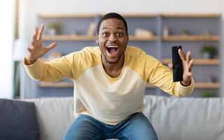 Happy african american man having fun at home, screaming with remote controller in his hand, watching football game on TV, empty space. Black guy enjoying his weekend, watching TV in living room photo
