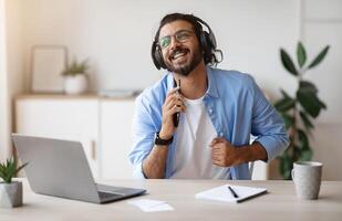 Cheerful indian freelancer guy singing and listening music with headphones while working with laptop at home office, using smartphone as microphone, enjoying his favorite song, copy space photo