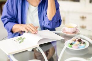 A retired woman in casual attire sits at a cafe table, reading a book while sipping tea. Colorful candies are placed beside her, creating a warm and inviting atmosphere. photo