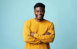 Cheerful handsome young african american guy in yellow sweater posing alone on blue studio background, holding hands crossed on chest and smiling at camera, copy space for advertisement photo