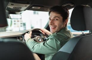 Handsome young man looking back at camera, sitting in car, parking auto, view from rear seat. Happy millennial guy buying automobile at modern dealership store, copy space photo