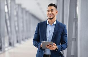 Portrait Of Happy Arab Man With Digital Tablet Standing At Airport Terminal, Handsome Middle Eastern Businessman Using Tab Computer While Waiting For Flight Departure, Enjoying Business Trip photo