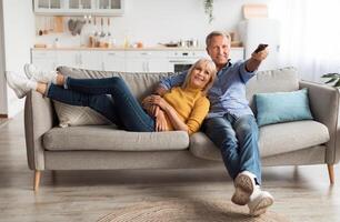 Relaxed Senior Couple Watching Television Together Sitting On Sofa And Pointing Remote Controller Switching Channels Relaxing At Home On Weekend. TV Programming Concept photo