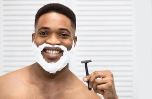 Smiling afro man with naked torso and shaving foam on his beard, holding razor, looking in mirror at bathroom. Comfort shaving concept, copy space photo