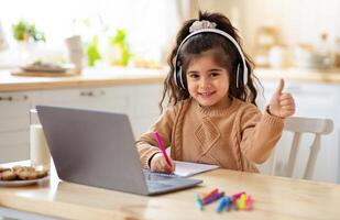 Distant Education. Adorable Little Girl In Headphones Using Laptop In Kitchen, Sitting At Table And Showing Thumb Up At Camera, Female Child Study Online With Computer And Drawing With Pencils photo