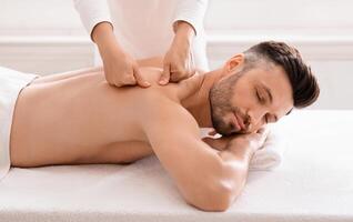 Middle aged bearded man attending modern male spa, laying on massage table, getting healing body procedure, enjoying his day at spa. Masseuse pressing on man back, using acupressure technique photo