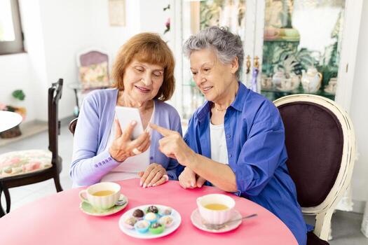 dos mayor mujer son sentado a un redondo mesa, compartiendo un alegre momento mientras mirando a un teléfono inteligente juntos. ellos disfrutar tazas de té y un plato de vistoso pasteles en un acogedor café ajuste. foto