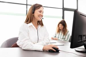 Two businesswomen are actively engaged in a corporate office setting. One is using a headset while operating a computer, and the other is writing notes. The atmosphere is productive and collaborative. photo