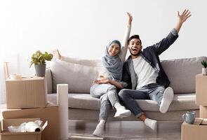 Emotional muslim family celebrating moving to their new house, sitting on new couch among paper boxes with stuff and having break while unpacking belongings, raising hands up and smiling, copy space photo