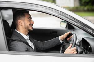 Happy middle eastern young businessman driving new auto, going to office, shot from outside, side view. Handsome successful arab guy manager going home after good day, looking at road and smiling photo