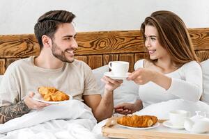 Romantic breakfast of couple in bed. Smiling attractive young woman holding tray with croissant and giving a cup of coffee to man with beard, free space photo