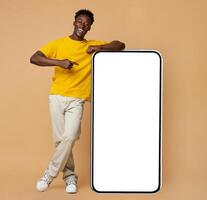 Happy Young Black Man Leaning And Pointing At Big Smartphone With White Blank Screen, Cheerful African Guy Showing Free Copy Space For Your Design, Standing Over Beige Background, Mockup Image photo