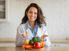 Portrait of cheerful dietitian in lab coat pointing at fresh fruits and vegetables on table, smiling at camera at clinic. Weight loss consultant recommending healthy plant based diet photo