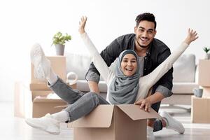 Positive muslim family playing with packing supplies while moving to their new house. Happy lady in hijab sitting in paper box and raising hands up, having fun with handsome husband while unpacking photo