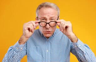 Poor Eyesight. Senior Man Can't See, Squinting Eyes Wearing Eyeglasses Having Problems With Vision, Looking At Camera, Yellow Orange Wall. Ophtalmic Issue In Older Age, Macular Degeneration Concept photo