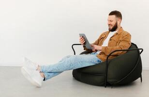 Side View Of Man Using Tablet Computer And Earbuds Earphones Watching Movie Online Sitting In Chair Over White Wall Indoors On Weekend. E-Learning, Internet And Technology Concept photo