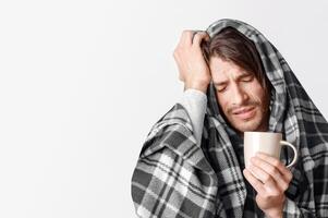 Treating flu and colds at home. Suffering man in blanket holds cup of hot drink and presses his hand to head isolated on gray background, studio shot photo