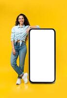 Full length of cheerful young lady standing near big smartphone with empty screen, demonstrating mockup for website, advertisement or app over yellow studio background photo