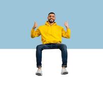 Happy young arab guy sitting on huge horizontal empty placard for advertisement over blue studio background, showing thumb up and smiling at camera, recommending something photo