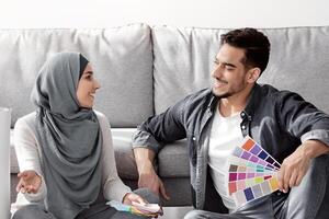 Creative arab family working on interior for their new house. Happy middle-eastern man and woman in hijab sitting on floor by paper boxes with color samples, choosing colors and textures, copy space photo