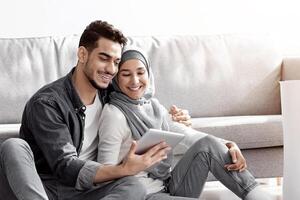 Happy muslim family sitting on floor in new apartment with digital tablet among boxes with belongings, buying furniture for house or working on design together, drinking tea and having conversation photo