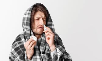Virus and cold. Man in blanket drips into his nose and holds napkin in his hand, face expresses suffering, isolated on gray background photo