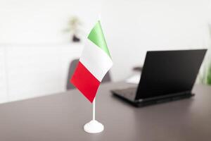 A small Italian flag is placed on a desk in an office environment next to a laptop. The setting suggests a professional atmosphere where work is being conducted. photo