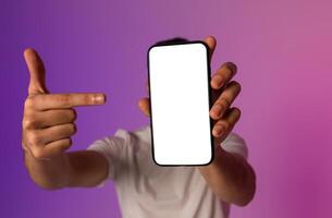 Unrecognizable young man holding smartphone in front of his face, pointing at empty screen, offering space for mobile app or website, demonstrating mockup for online ad in neon light, selective focus photo