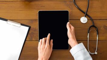 Modern private practice of doctor and patient visit. Hands of female therapist in white coat works on tablet with blank screen on wooden table with stethoscope, apple, plant and glasses, top view, pov photo