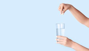 Female hands dropping pill into glass of water on light background, close up. Blank space. Panorama photo