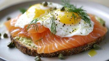 A close-up image of a plate of smoked salmon and avocado toast topped with fried eggs, garnished with dill and capers. The toast is sliced thick and has a slightly toasted crust. photo