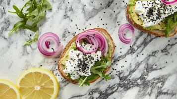 A close-up of a plate with two slices of avocado toast. Each slice is topped with ricotta cheese, red onion, arugula, and black sesame seeds. Lemon slices are visible at the bottom of the image. photo