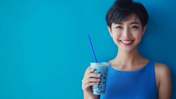 A young woman with short hair joyfully holds a blue drink with bubbles while standing in front of a bright blue wall. Her cheerful expression highlights the refreshing nature of her beverage. photo
