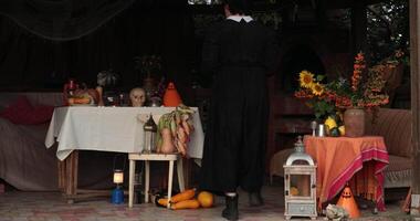 Woman witch in nun costume sets table Halloween decorations with mystery props. Goth witch preparing Halloween cupcakes treats in rustic gazebo with retro lanterns and fall arrangements video