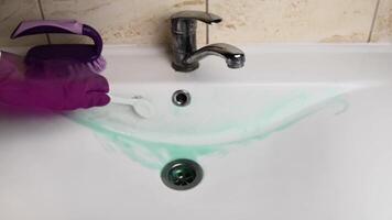 Time lapse female hands in purple gloves scrub and clean white ceramic sink with green cleaning liquid and brushes, then flow out with water from opened chrome faucet. Housework chore concept video
