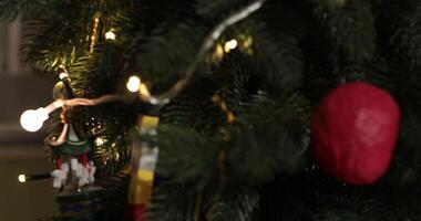 Soft focus to artificial Christmas tree decorated with retro Christmas ornaments and golden lamp garland in night. Wooden nutcracker toy on traditional Christmas tree. Rack focus closeup video