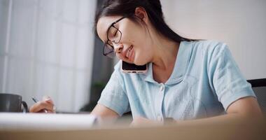 Handheld selective focus shot, Female freelancer sitting at desk working and talking on the mobile phone at the same time in home office. Working from home video