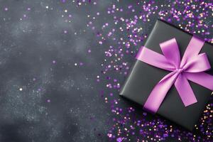 Purple bow on black gift box with glittering confetti scattered on dark surface photo