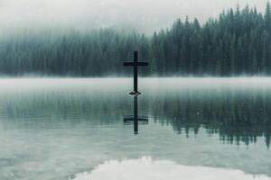 Reflection of a cross in calm water surrounded by mountains during early morning mist photo