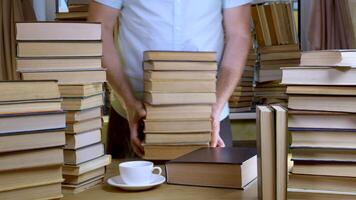 A bearded man reads a book in the library. Stacks of books and cup of tea on table. Concept of Reading and Education. video