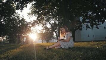 A girl is reading a book in the sunset rays, sitting on the grass in the park. video