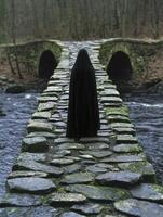 Mysterious figure in dark cloak on mossy stone bridge photo